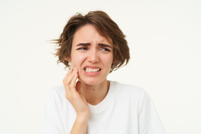Close up of woman has a toothache, touches her teeth and frowns from painful discomfort, stands over white background
